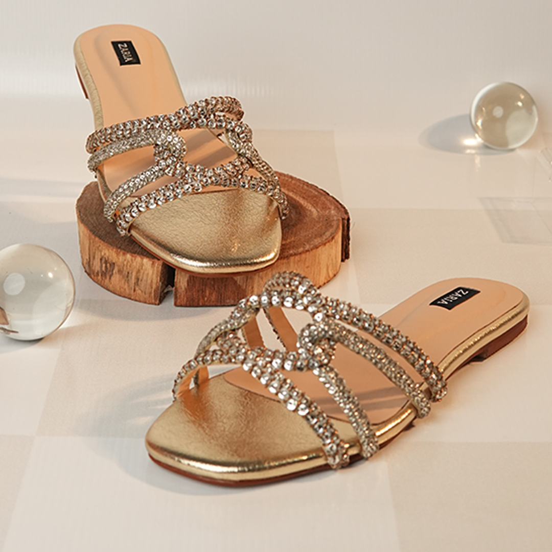 Zaria ZARAH flat sandals with crystal-covered straps and soft gold footbeds, displayed beside a wooden stand.